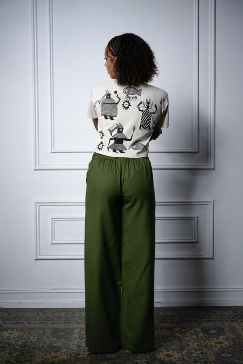 A female model stands with her back to the camera wearing a cream-colored short-sleeve crop top with a black African graphic print, Cooyah logo, and olive green wide-leg trousers, set against a white paneled wall.