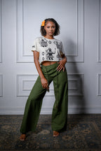 A model stands in a room with white paneled walls and a patterned rug, wearing olive green wide-leg pants and a beige cropped graphic t-shirt with black African illustrations.