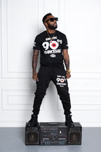 Man wearing a black t-shirt and pants with '90s Dancehall' text, standing on a boombox against a white wall. Cooyah - Jamaican owned clothing brand since 1987.