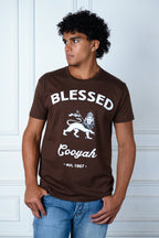 Man wearing a brown t-shirt with 'Blessed Cooyah' text and lion graphic against a white paneled wall.