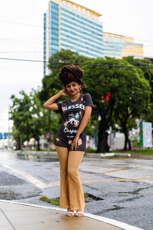 Model posing on a city street in Trinidad with modern buildings in the background. She has beautiful locs styled like a crown and is wearing a shirt with Blessed text on the fornt.