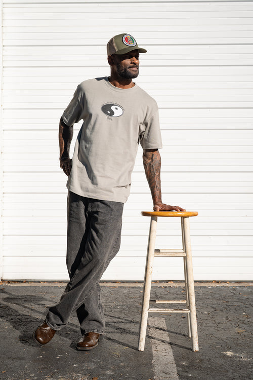 Front view of man wearing a gray Cooyah t-shirt with a Yin Yang design and cap, standing next to a wooden stool against a white wall.