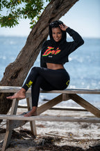 Woman sitting on a wooden bench by the ocean wearing a cropped black hoodie with colorful text "Irie" in rasta colors and leggings. Complete with Cooyah logo.