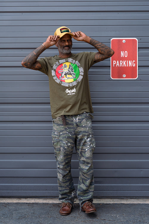 Man wearing an olive green Cooyah t-shirt with a colorful Lion of Judah rasta graphic and camouflage pants, standing against a gray background. Reggae Massive trucker hat.