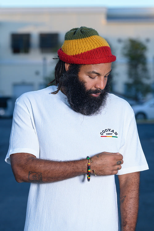 Man wearing a colorful, rasta beanie and white gauze shirt with "Cooyah" embroidered text, standing outdoors.