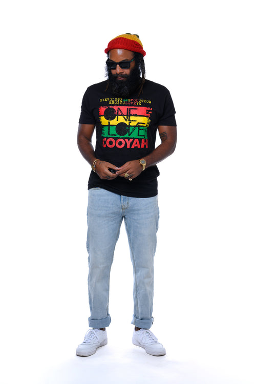Reggae musician Lan Lawd wearing a black t-shirt with colorful "One Love" text, light blue jeans, and a red, yellow, and green beanie on a white background.  Cooyah, the official reggae clothing brand since 1987.