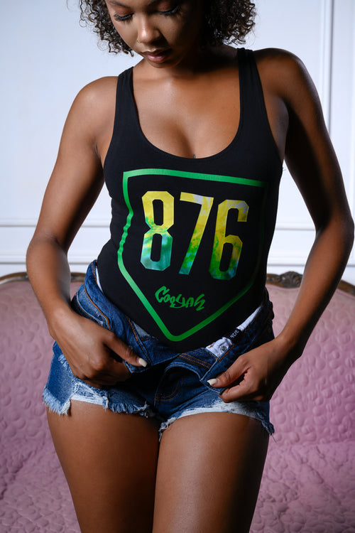 A woman wearing a black bodysuit with a  876 graphic in yellow-green gradient and Cooyah logo paired with denim shorts, standing in front of a pink textured background.