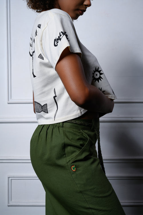 A model shown from the side and back wearing a cropped t-shirt with black Afrian prints, Cooyah logo, and olive green pants, standing against a white paneled wall.