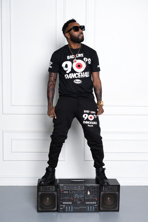 Man wearing a black t-shirt and pants with '90s Dancehall' text, standing on a boombox against a white wall.  Cooyah - Jamaican owned clothing brand since 1987.