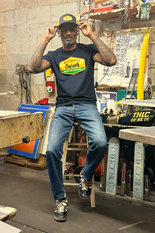 Male model wearing a Cooyah t-shirt and cap with 'Best Quality Made in Jamaica" text in a workshop setting.