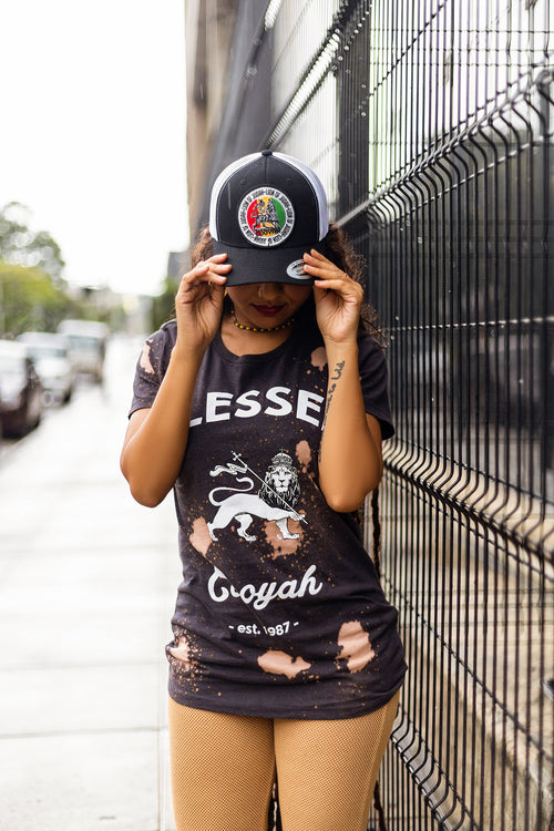 Model wearing a black distressed t-shirt with Lion graphics and Blessed text, holding a cap above her head, standing against a fence.  Cooyah Clothing Brand.