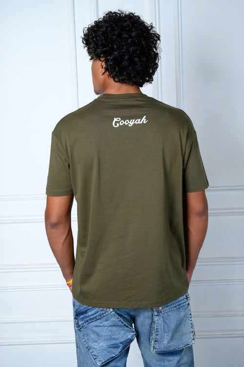 Model wearing an olive green t-shirt with 'Cooyah' logo on the back, standing against a white wall.