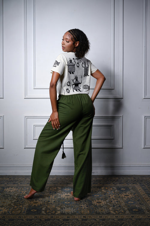 Cooyah Clothing - A model stands barefoot in a room with white paneled walls and a patterned rug, wearing olive green wide-leg pants and a beige crop top with African graphics.