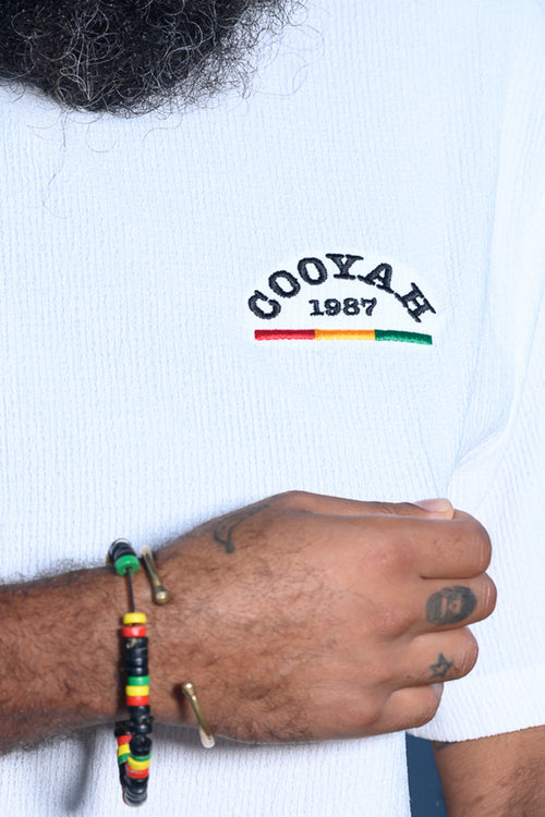 Man wearing a white t-shirt with 'COOYAH 1987' text, holding a rasta-colored bracelet. 