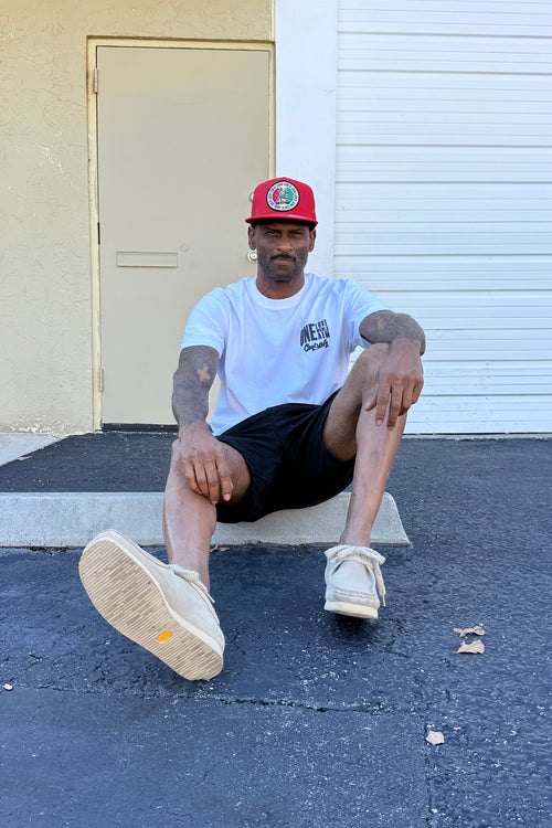 Man sitting on a step wearing a white t-shirt, black shorts, and a red cap.  Cooyah Clothing Brsnd.