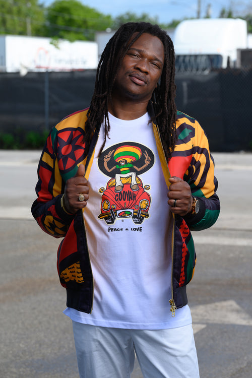 Reggae singer Kumar Fyah wearing a Cooyah graphic tee with a colorful red, yellow, and green VW Beetle car design and Rasta man cartoon graphic layered under a matching multicolor patterned bomber jacket. He is standing outdoors on a paved surface with a fence and trees in the background.