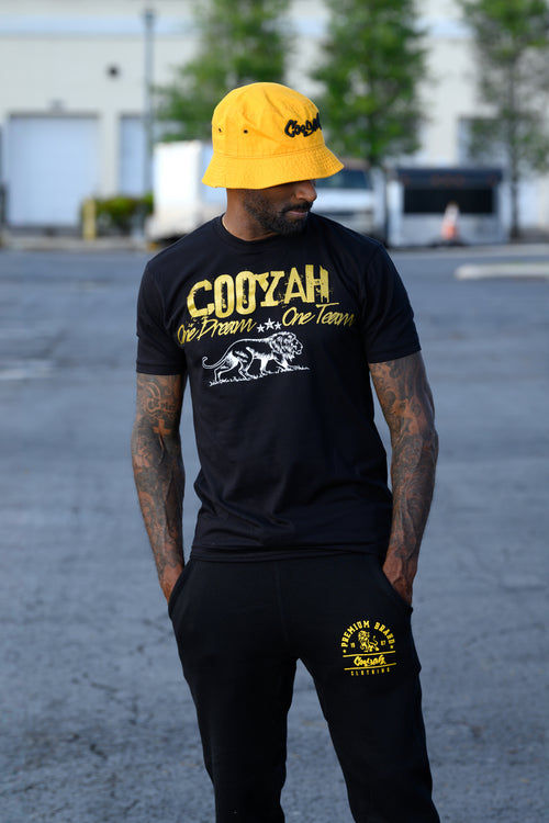 Cooyah Jamaica. One Dream, One Team, men's graphic tee in black with metallic gold lettering and white lion print.  As a Jamaican owned clothing brand since 1987, we take pride in bringing you this classic piece. Don't wait, get yours now!