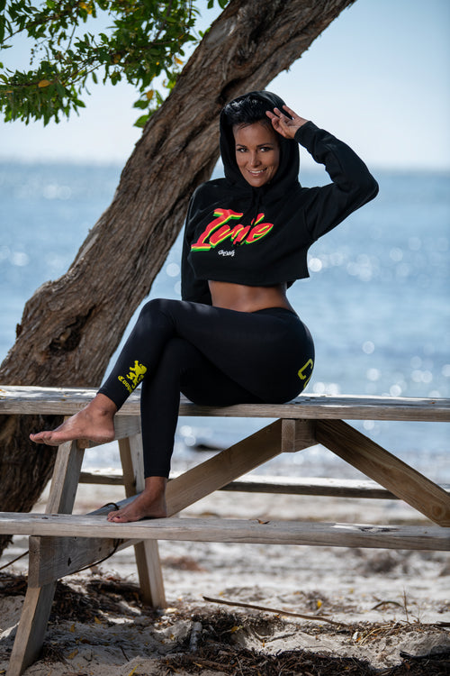 Woman sitting on a wooden bench by the ocean wearing a cropped black hoodie with colorful text "Irie" in rasta colors and leggings.  Complete with Cooyah logo.