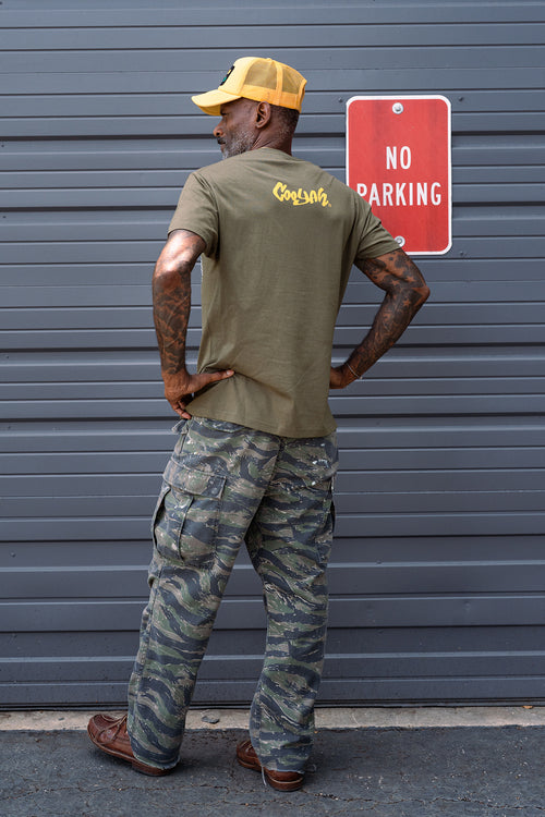 Man wearing a green Cooyah t-shirt with yellow text and camouflage pants standing next to a 'No Parking' sign.