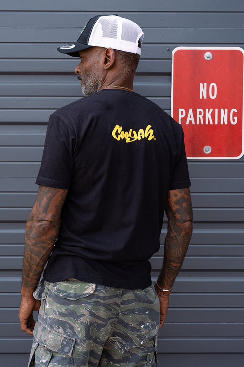 BAck view of man wearing a black Cooyah t-shirt with a colorful Lion of Judah rasta graphic and camouflage pants, standing against a gray background.