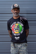 Man wearing a black Cooyah t-shirt with a colorful Lion of Judah rasta graphic and camouflage pants, standing against a gray background.