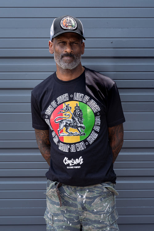 Man wearing a black Cooyah t-shirt with a colorful Lion of Judah rasta graphic and camouflage pants, standing against a gray background.