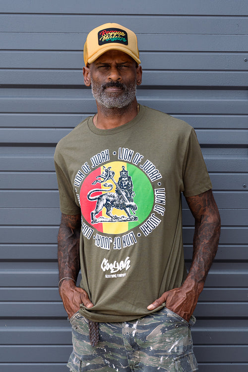 Man wearing a green short sleeve t-shirt with a colorful Lion of Judah graphic and a yellow embroidered cap with Reggae Massive patch, standing against a gray metallic background.  We are Cooyah the offiical reggae clothing brand since 1987.