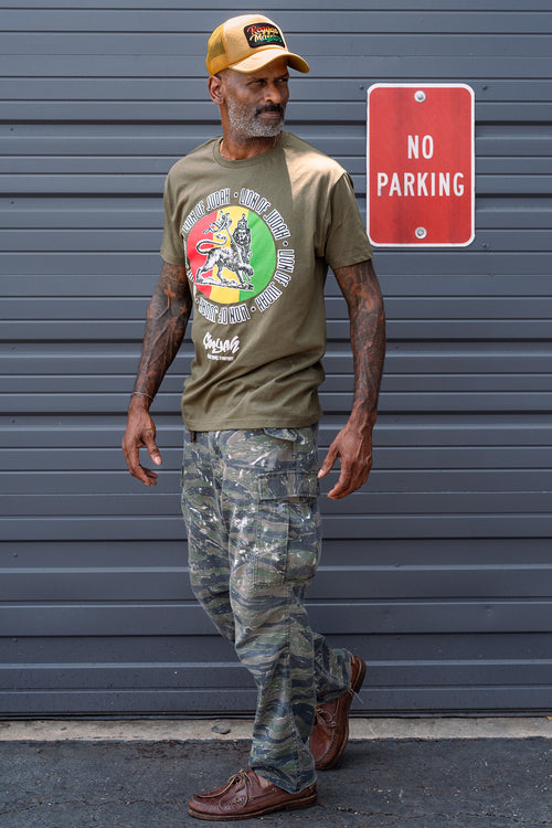 Man wearing an olive green Cooyah t-shirt with a colorful Lion of Judah rasta graphic and camouflage pants, standing against a gray background.  Reggae Massive trucker hat.
