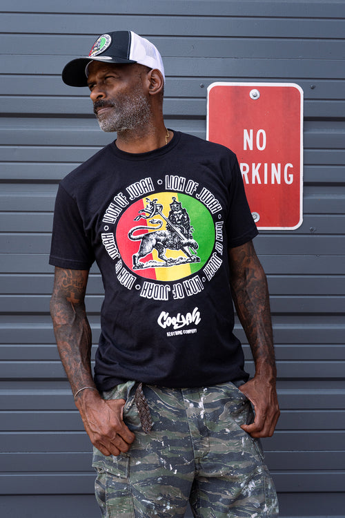 Man wearing a black Cooyah t-shirt with a colorful Lion of Judah rasta graphic and camouflage pants, standing against a gray background.