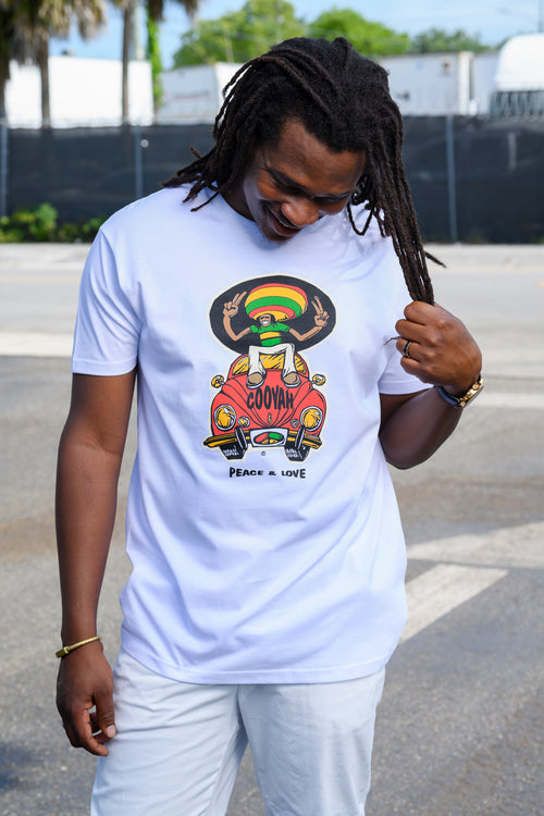 A man with dread locks wearing a white graphic t-shirt with a red, gold, and green cartoon illustration of a rasta man riding a red Volkswagon Beele with 'COOYAH' written on it and 'PEACE AND LOVE' below, standing outdoors on a paved surface.