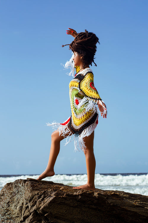 Cooyah empress ii-kaya wearing a colorful, patterned dress with fringe standing on a rock with ocean and sky in the background.  