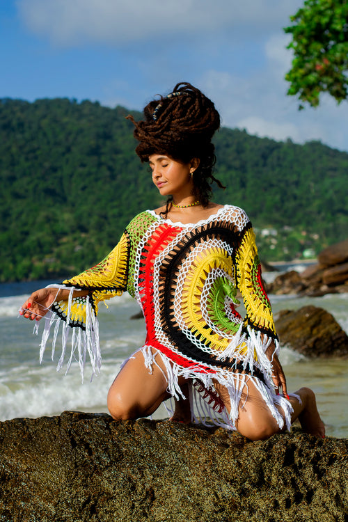 Cooyah Jamaica - Rasta woman in a colorful crochet dress sitting on a rock by the ocean with a mountainous background.